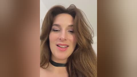 Media: Video of a fair-skinned woman with long, wavy brown hair, wearing a black choker necklace, smiling, and slightly tilting her head. Background is a blurred, neutral-toned gradient.