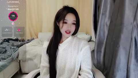 Media: Video of an East Asian woman with long, straight black hair, wearing a white, textured knit sweater, seated in a cozy, dimly lit room with a bed and gray curtains in the background.