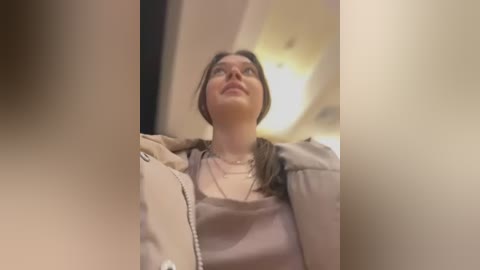 Media: Video of a young woman with fair skin and brown hair, wearing a beige jacket and blouse, gazing upward in a dimly lit airplane cabin.