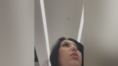 Media: Video of a young woman with long black hair, fair skin, and full lips, looking up, against a modern, white ceiling with rectangular lighting fixtures.