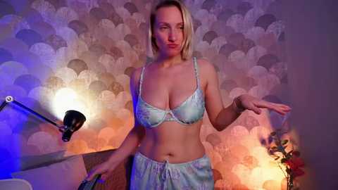 Video of a blonde Caucasian woman with fair skin wearing light blue lingerie, standing in a dimly lit room with a patterned wallpaper and a desk lamp.