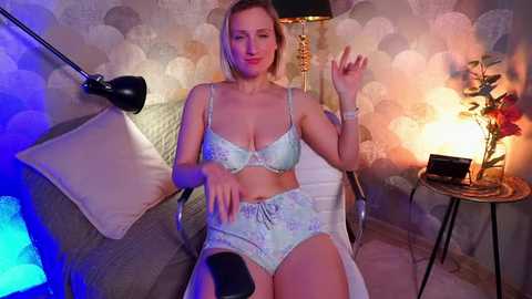 Media: A video of a blonde Caucasian woman in light blue lingerie, seated on a chair in a dimly lit room with patterned wallpaper, a lamp, and a camera.