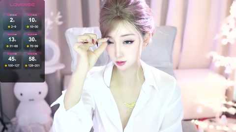 Media: Video of a young East Asian woman in a white blouse, adjusting her eyeglasses, in a softly lit, pastel-themed room with teddy bears and flowers.