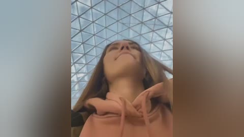 Video of a young woman with light skin, long brown hair, and closed eyes, adjusting her pink hoodie, viewed from a low angle, with a geometric glass ceiling in the background.