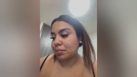 Media: Video of a Latina woman with long brown hair, wearing black lingerie, standing in a bathroom with a white ceiling light and beige walls.