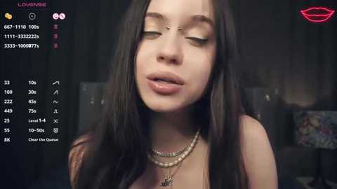 Media: Video of a young woman with long dark hair, fair skin, and a nose ring, wearing multiple gold necklaces, looking seductively into the camera. Background is dark and blurred, with a virtual lipstick overlay.