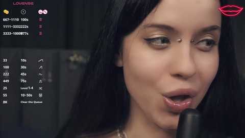 A close-up video of a woman with long black hair, wearing makeup and a silver nose ring, singing into a microphone.