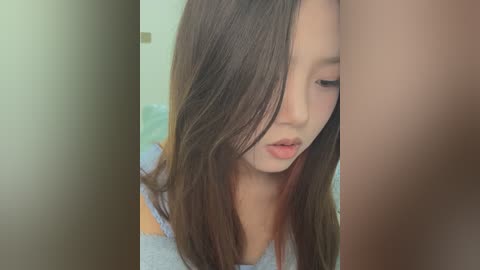 Media: Video of a young Asian woman with long, straight brown hair, wearing a gray sweater, looking down with a neutral expression. Background features a blurred, light-colored wall.