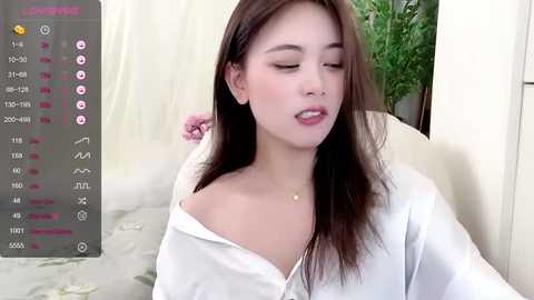 Media: A video of an East Asian woman with long brown hair, wearing a white off-shoulder top, and a small necklace, sitting indoors with a green plant in the background.