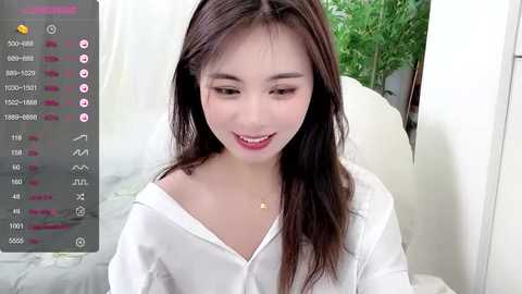 Media: A video of an Asian woman with long dark hair, fair skin, and light makeup, wearing a white off-shoulder top, smiling. The background includes a white bed and a green plant.