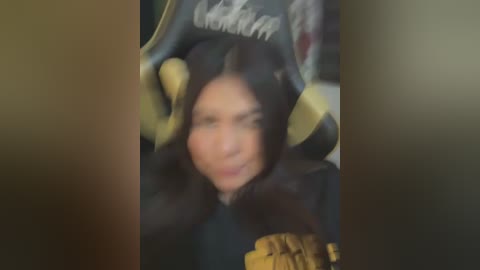 Media: A blurry video of a person with dark hair, wearing a black jacket, seated in a car with visible yellow seatbelts.