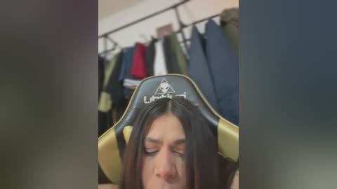 Media: Video of a woman wearing a black and yellow gaming chair with a triangle emblem, her long dark hair covering her face. Clothes hang on a rack in the background.