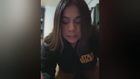 Video of a young woman with shoulder-length brown hair, wearing a black \"Star Wars\" T-shirt, leaning into a dimly lit doorway, looking down.