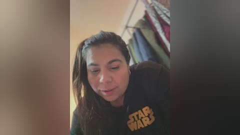 Media: Video of a young woman with long brown hair, wearing a black \"Star Wars\" hoodie, smiling softly, in a dimly lit room with a hanging clothes rack in the background.