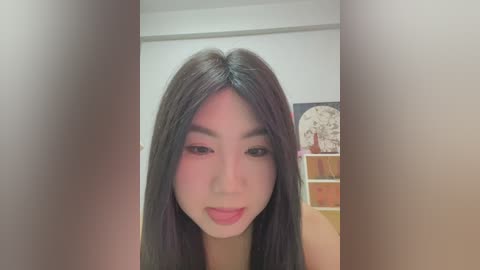 Media: A video of a young East Asian woman with long, straight black hair, fair skin, and pink lipstick, looking directly at the camera. She's in a modern room with a white wall, a wooden shelf, and a decorative clock in the background.