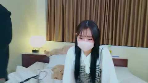 Media: A video shows a young Asian woman with long black hair and a surgical mask kneeling on a bed, wearing a white and black plaid dress, in a simple bedroom with beige curtains.