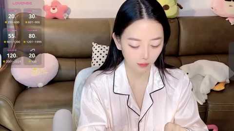 Media: Video of a young Asian woman with straight black hair, wearing a white blouse, sitting on a beige leather couch. Background includes stuffed animals and a digital overlay showing live stats.