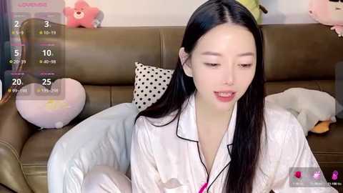 Media: Video of a young Asian woman with long black hair, wearing a white bathrobe, sitting on a brown leather couch, surrounded by plush toys, including a large pink bear.