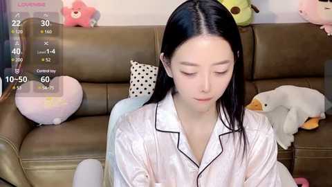 Media: Video of a young Asian woman with long black hair, wearing a white satin pajama top, sitting on a beige couch. Background includes stuffed animals and a plush pillow.