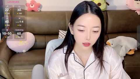 Media: Video of an Asian woman with long black hair, wearing a white shirt, sitting on a brown leather couch in a cozy room with stuffed toys and a calendar overlay.