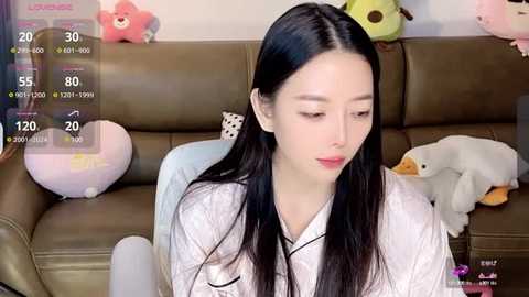 Media: Video of an East Asian woman with long black hair, wearing a white robe, seated on a brown leather sofa. Background features stuffed toys, including a panda, a bunny, and a white dog. Live streaming overlay with viewer counts and chat messages.