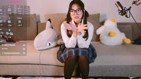 Media: Video of an Asian woman with glasses, dressed in a white shirt, plaid skirt, and black tights, sitting on a beige sofa with plush toys, holding a phone.