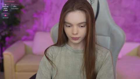 A video of a young, fair-skinned, long-haired, androgynous person sitting in a gaming chair, with a pink brick wall background and a blurred plant in the left corner.