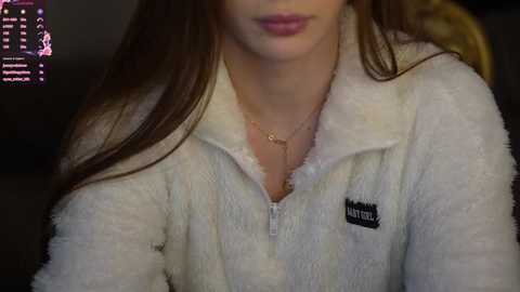 Media: Video of a young woman with light skin and long brown hair, wearing a white, fuzzy zip-up hoodie with a small logo on the chest. She has a thin gold necklace. The background is dark and blurred, with a colorful digital screen partially visible.