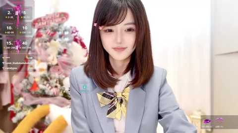 Media: A video of an East Asian woman with straight, shoulder-length dark hair, wearing a light blue blazer and yellow bow tie, standing in a festive, brightly lit room with a decorated Christmas tree in the background.