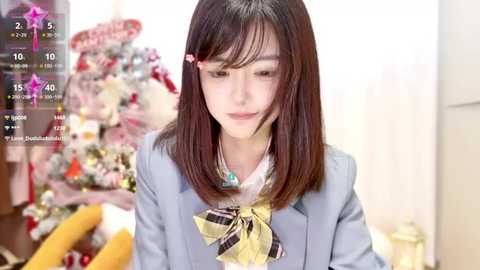 Media: Video of an Asian woman with straight, shoulder-length dark hair, wearing a light blue blazer with a yellow bowtie, standing in a bright, festive room with a decorated Christmas tree in the background.