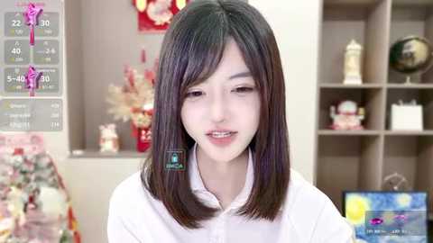 Media: Video of a young Asian woman with shoulder-length black hair, wearing a white blouse, smiling, in a cozy room with a TV showing a weather forecast and bookshelves.