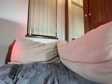 Video of a bed with a beige pillow and blue duvet, illuminated by a soft pink light, set against a wooden door and window with white blinds.