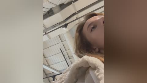 Media: Video of a young woman with fair skin and blonde hair, wearing a beige sweater, standing in a warehouse with white panels and metal beams in the background.