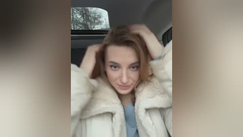 Media: Video of a young woman with light skin and light brown hair, wearing a fluffy white coat and blue top, sitting in a car with bare trees outside the window.