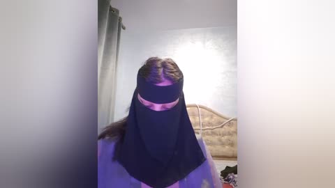 Media: Video of a person wearing a black niqab in a dimly lit room with beige walls, a bed, and purple lighting.