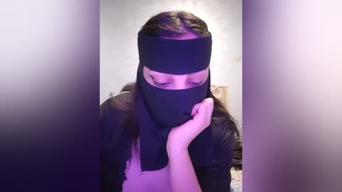 Media: Video of a woman with long brown hair, wearing a black mask and a black top, with her hands covering her face, standing in a dimly lit room.