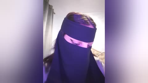 Media: Video of a person with long brown hair wearing a black niqab, partially revealing their eyes, standing in a room with beige curtains and white walls, bathed in purple lighting.