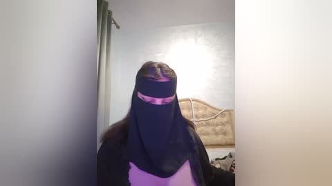 Media: A video of a person wearing a black burqa, standing in a dimly lit room with white walls and a beige headboard.