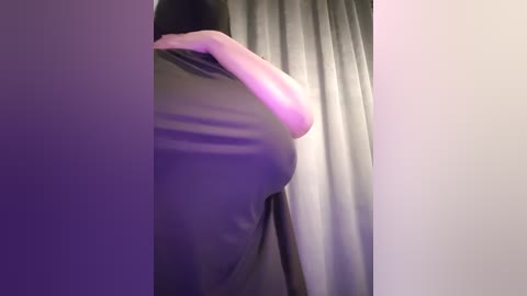 Media: Video of a person's backside, wearing a black dress, standing in front of a purple and grey curtain, with a purple light illuminating the scene.