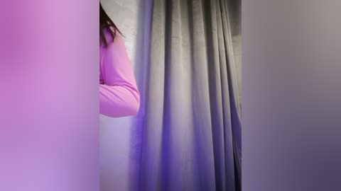 Media: A video of a person's arm in a light pink long-sleeve shirt peeking from behind a grey curtain, illuminated by soft, purple lighting.