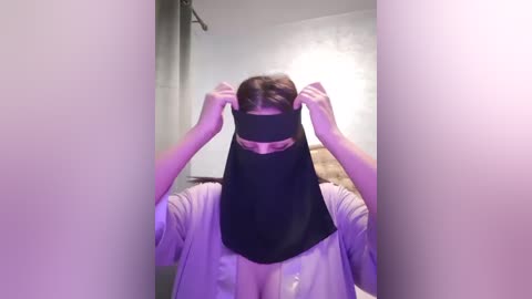 Media: A video of a person with a black face mask, adjusting it, wearing a light-colored shirt, in a dimly lit, industrial-style room with concrete walls and a faint purple light.