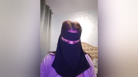 Media: Video of a person with long red hair, wearing a black niqab and white shirt, standing in a room with beige walls, a bed, and gray curtains.