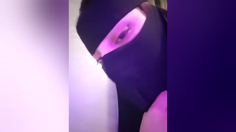 Media: Video of a person wearing a black mask with a purple tint, partially obscured, set against a blurred, gradient background of purple and white. The image is low-resolution and grainy, with a focus on the face and mask.