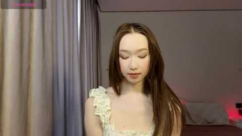 A video of a young, fair-skinned Asian woman with long brown hair, wearing a white lace dress, standing in a dimly lit bedroom with soft, pastel-colored walls and a bed with white sheets.