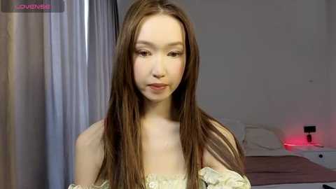 Media: Video of a young East Asian woman with long, straight brown hair, wearing a beige off-shoulder dress, in a minimalist bedroom with white walls, a bed, and a red nightlight.