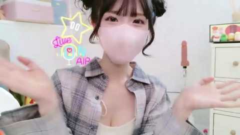 Media: A video of an East Asian woman with light skin, dark hair, and bangs, wearing a face mask, gray plaid shirt, and a white top, posing playfully indoors with colorful, starry virtual overlay.