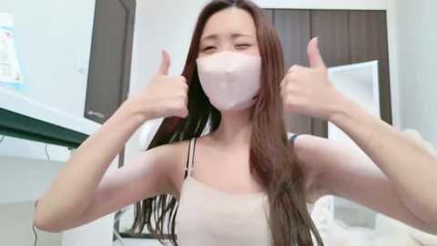 Media: Video of an Asian woman with long brown hair, wearing a white face mask, giving thumbs-up in a modern, bright room with white walls and dark wooden furniture.