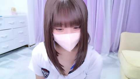 Media: Video of a young Asian woman with straight brown hair, wearing a white mask, white shirt, and blue bra, seated indoors near a white dresser and light purple curtains.