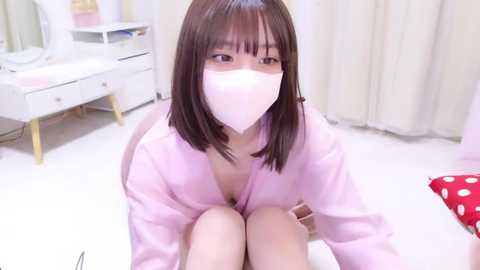 A video of an Asian woman with straight, shoulder-length brown hair, wearing a pink robe, sitting on a white floor in a brightly lit room with white furniture and curtains.