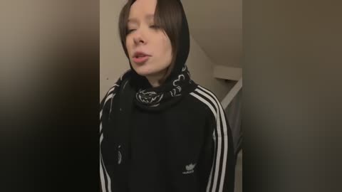 Media: Video of a pale-skinned, androgynous person with shoulder-length brown hair, wearing a black Adidas tracksuit with white stripes, and a black hooded sweatshirt featuring a panda design. The background is a dimly lit, beige hallway.